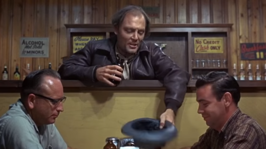 A man in a bar with his hat held out to two seated patrons. It appears he's trying to get some money.