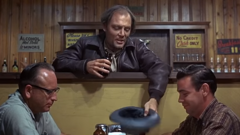 A man in a bar with his hat held out to two seated patrons. It appears he's trying to get some money.